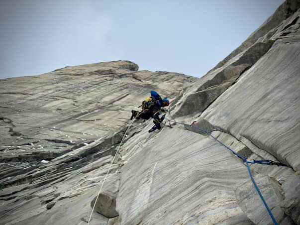 A climber in blue jacket with ropes trailing from his harness takes a moment to pause to navigate steep terrain