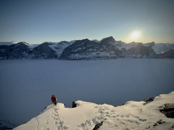 A climber at the top of the route, with a snow covered summit ahead and a frozen fiord below in the background