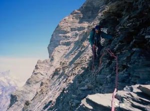 A roped-up climber traverses an exposed area of loose rock.