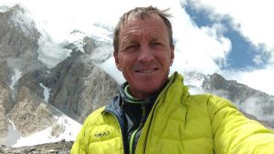 Denis Urubko smiles with a massive mountain face (Broad Peak?) behind.