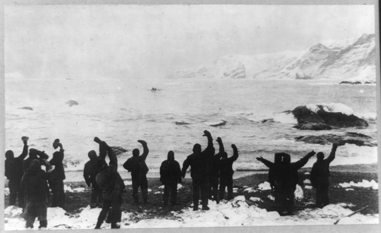 A group of people stand on a rocky shore waving at a small boat in the distance.