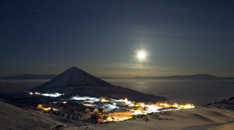 A view of a small cluster of buildings below a cone-shaped hill, with a dark sky and the Moon shining.