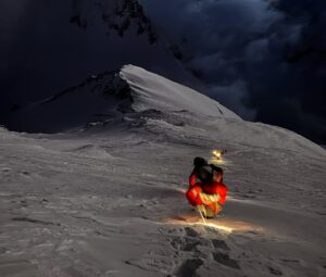 A climber with a headlamp on a snow ramp, clouds below him.