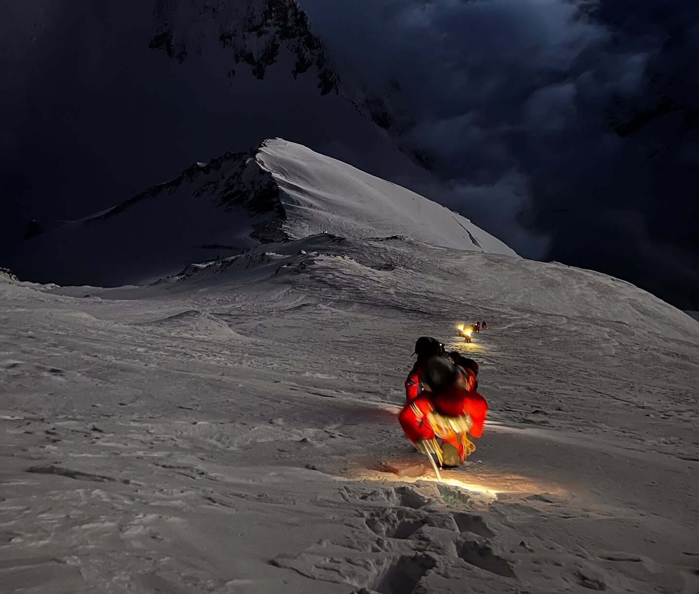 A climber with a headlamp on a snow ramp, clouds below him.