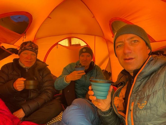 The climbers hold plastic cups inside a yellow tent. 