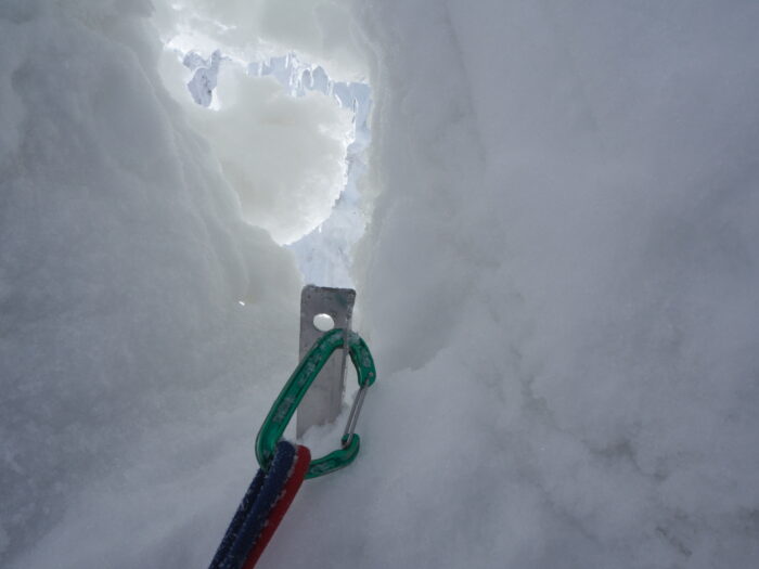 A tunnel-like hole in the ice, with a snow anchor and a carabiner.