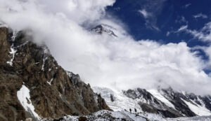 K2 wrapped in clouds and two climbers walking towards it from the glacier at the base.