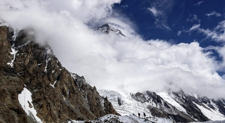 K2 wrapped in clouds and two climbers walking towards it from the glacier at the base.