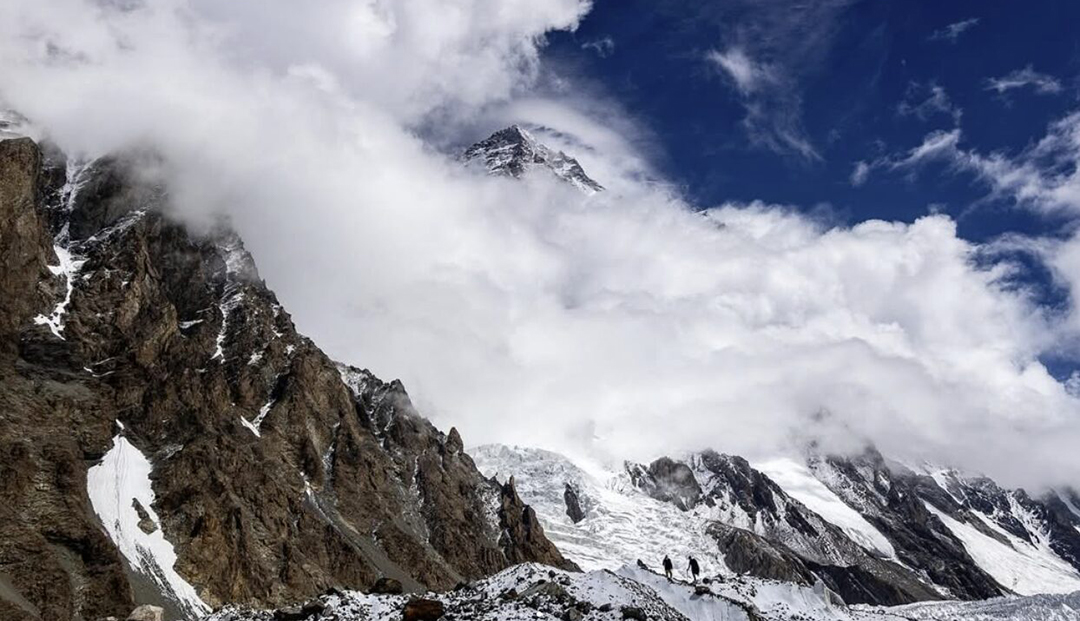 K2 wrapped in clouds and two climbers walking towards it from the glacier at the base.