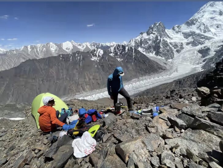 Two climbers in front of a small tent pitched on rocky morraine terrain.