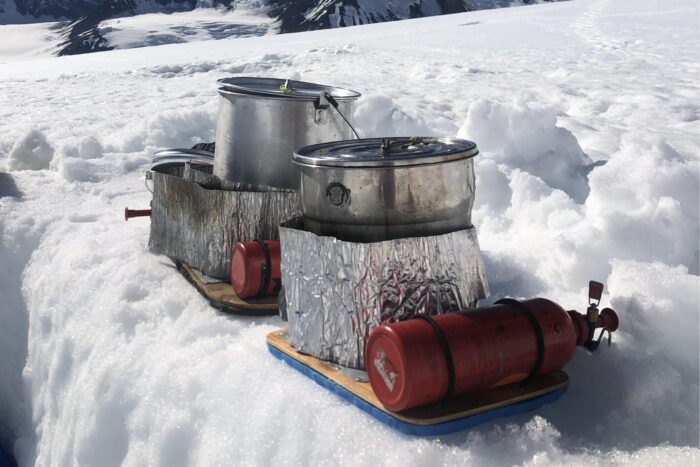 Two whisperlites are in the snow, with windscreens and large pots on top of them. 