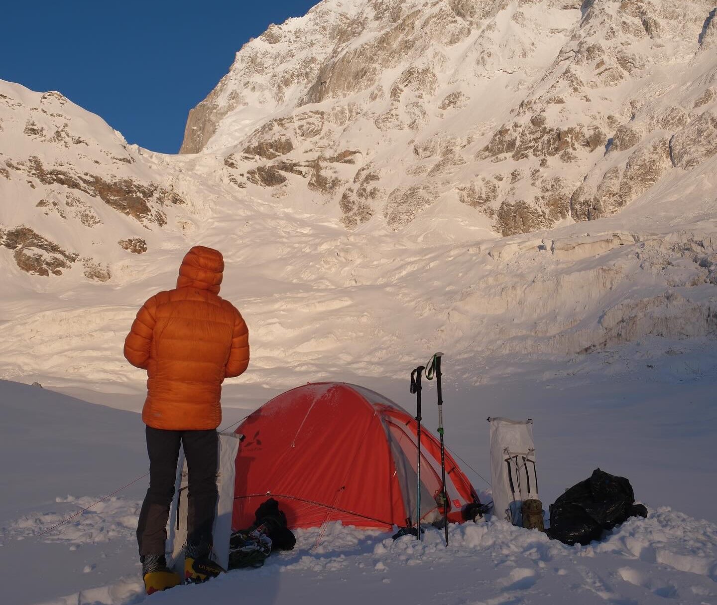 A climber looking up a moutnain face from behind his tent on a snowy plane.