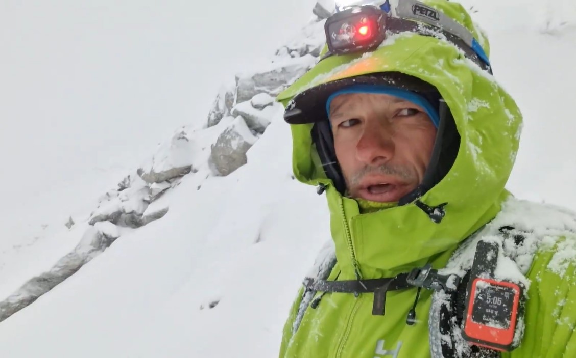 Video frame showing Kowalewski in a snowy day on a snow slope, a headlamp and hood on his gore-tex jacket.
